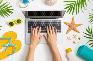 Hands typing on a laptop surrounded by summer-themed items, including yellow polka dot flip-flops, a starfish, sunglasses, and tropical leaves, evoking a fun and relaxed work-from-anywhere atmosphere