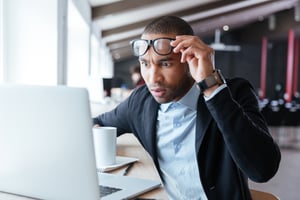 A surprised man lifting his glasses while looking at a laptop screen with a cup of coffee beside him.