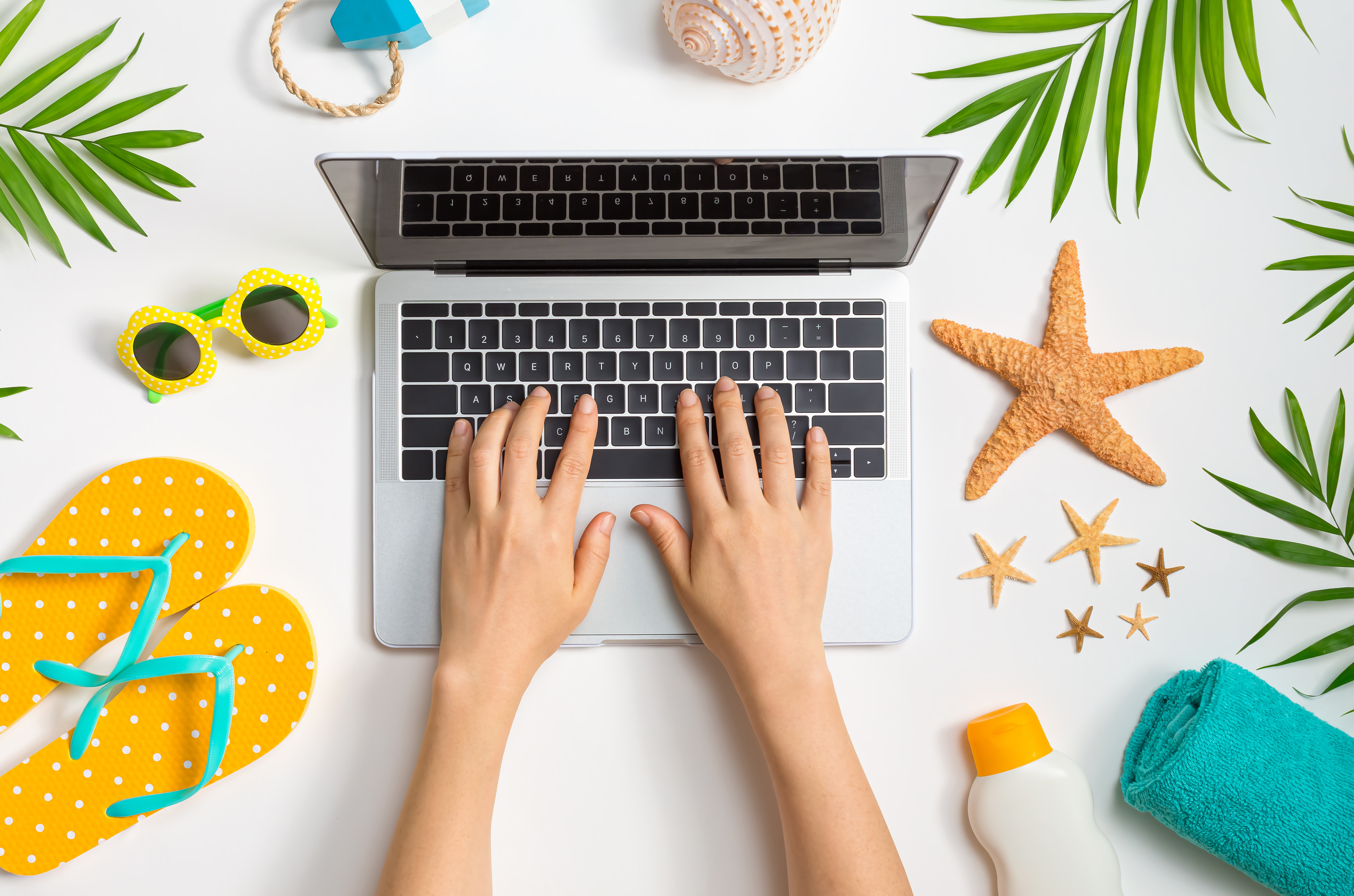 Hands typing on a laptop surrounded by summer-themed items, including yellow polka dot flip-flops, a starfish, sunglasses, and tropical leaves, evoking a fun and relaxed work-from-anywhere atmosphere