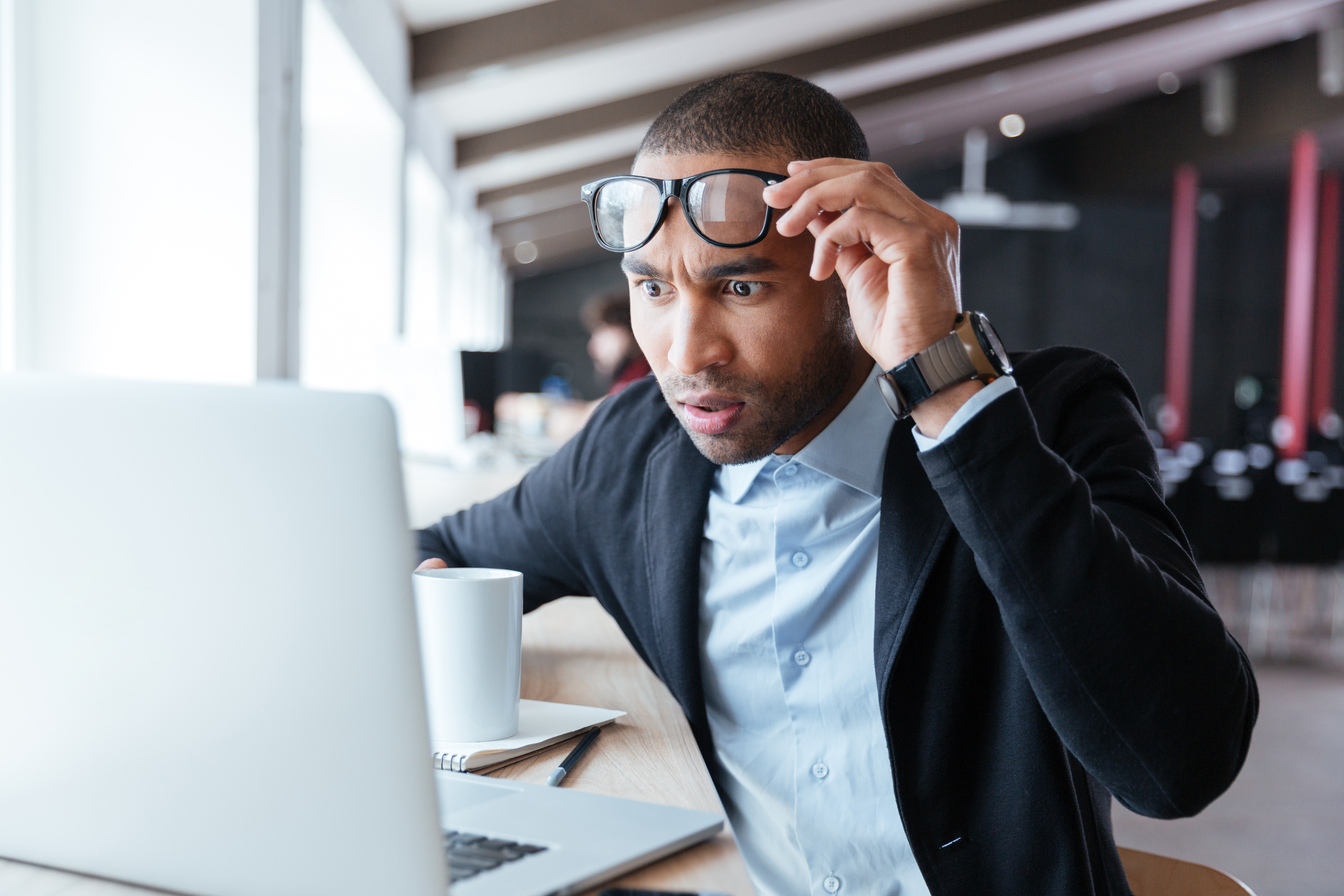 A surprised man lifting his glasses while looking at a laptop screen with a cup of coffee beside him.